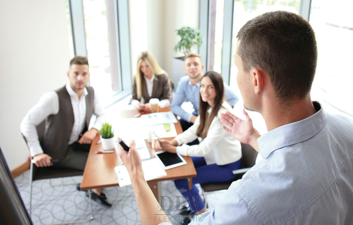 Eine Gruppe von fünf Personen sitzt um einen Tisch in einem hellen Raum. Eine Person steht, spricht und gestikuliert. Die anderen hören aufmerksam zu, Laptops und Unterlagen sind auf dem Tisch.
