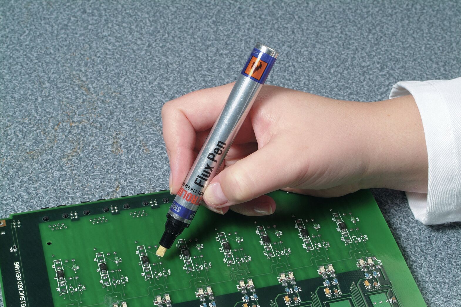 A hand holds a marker over a green circuit board. The focus is on the marker, which is yellow, while the circuit board displays numerous electronic components.