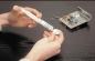 Preview: A person's hands hold a small plastic body while they align a syringe with a silvery substance on it. In the background, there is an electronic component with many small parts.