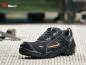 Preview: The image shows a black work shoe with a round toe cap. It has orange accents and a non-slip sole. The shoe is placed on a gray, textured surface.