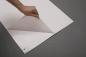 Preview: In the picture, you see a hand lifting a corner of a white sheet. Beneath it, a smooth, transparent film is visible. The background is gray.