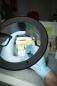 Preview: A hand with blue gloves is holding a dental model under a magnifying glass. The model displays white teeth and a jaw. In the background, work tools and a blurred environment can be seen.