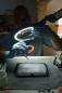 Preview: A person wearing glasses and gloves is working under a lamp. They are holding a model of a denture in their hand while working on it with a tool. The background is blurred.