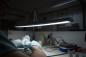 Preview: Auf dem Bild sieht man einen Arbeitstisch mit einer hellen Lampe. Eine Hand mit blauen Handschuhen hält etwas Kleines. Um den Tisch liegen verschiedene Utensilien und Materialien.