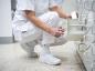 Preview: Eine Person in weißer Kleidung und weißen Schuhen sitzt auf dem Boden und greift in ein Regal, um etwas herauszuholen. Der Boden ist hell und die Umgebung wirkt sauber und ordentlich.