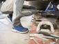 Preview: The image shows a person in work clothes, sitting at knee height in front of a car. They are wearing blue shoes and working with tools on a workbench, surrounded by car parts.