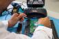 Preview: A person in a white coat is soldering with a soldering iron on a green circuit board. In the background, a Weller device is visible. The table is light blue.