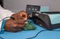 Preview: A hand works on a green circuit board, surrounded by technical devices. A device in light blue stands next to it, used for maintenance or testing.
