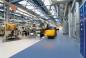 Preview: The image shows a modern factory hall with high ceilings, blue steel beams, and a bright floor. On the left side are machines and shelves, while a forklift is on the move.
