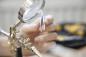 Preview: Auf dem Bild ist eine Hand zu sehen, die eine feine Pinzette hält. Ein vergrößerndes Glas ist an einem Halter befestigt. Im Hintergrund sind Werkzeuge sichtbar. Die Atmosphäre ist handwerklich.