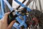 Preview: The image shows a hand holding a tool while working on the rear brake of a blue bicycle. The brake features a disc and sprockets are visible.