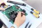 Preview: Auf dem Bild sieht man eine händige Montage oder Reparatur einer grünen Leiterplatte mit vielen elektronischen Bauteilen. Nebenan liegen Schraubenzieher und Zangen.