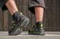 Preview: In the picture, you can see the feet of a person in sporty, black shoes with yellow accents. The pants are short and the background consists of a wooden structure.