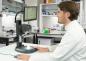 Preview: A man in a lab coat is sitting at a table, looking at a monitor connected to a microscope. In the background, shelves with laboratory utensils are visible.