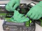 Preview: In the picture, there are two hands wearing green disposable gloves, taking another glove out of a package. Several packages are visible in the background.