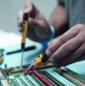 Preview: A person is working on a green circuit board. They are holding a soldering iron in their right hand and using tweezers in their left hand. The board has colorful components.
