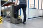 Preview: A man stands on a machine base with one foot. The machine is gray and is located in a warehouse with shelves and a white wall in the background. Safety markings are visible.