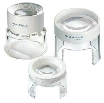 The image shows a magnifying glass in a compact, round design. At the top is a clear lens insert, below which is a slightly curved base made of transparent plastic. The surface is predominantly white.