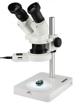The image shows a microscope with two eyepieces, a stable stand, and an illuminated base. On it lies a small object for viewing. It is predominantly white.
