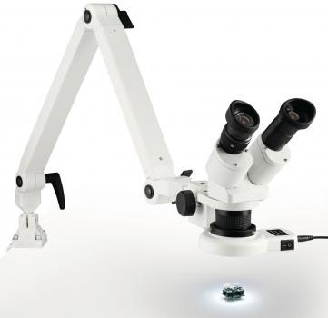 The image shows a white microscope with two eyepieces and a movable arm. Below it, an illuminated object, probably a tiny detail, is clearly visible.