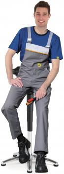 In the picture, a smiling young man is sitting on a stool. He is wearing a blue T-shirt and grey overalls. His black shoes are on the floor, and he is casually resting one hand on his knee.