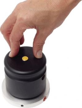 A hand presses on a round, black button, which is located on a cylindrical housing. The button has a small yellow dot in the center. The housing is on a gray, round base.