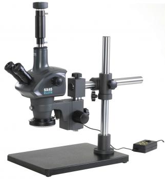 The image shows a microscope with a stable base. It has two eyepieces, an elongated objective, and a rotatable stage. A cable runs from the base. It is in dark gray.