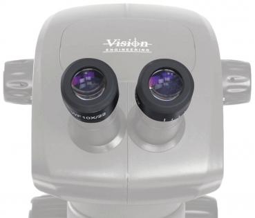 The image shows a binocular microscope with two large, round eyepieces. The surface is light gray with the 