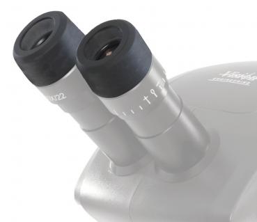 The image shows the eyepieces of a microscope. They are silver with black, rubberized edges. A scale indicates the magnification. The rest of the microscope is blurry in the background.