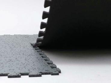 The image shows a black, textured surface made of thick mats stacked at one edge. One mat is slightly lifted, while the other is flat. The surface has a rough texture.