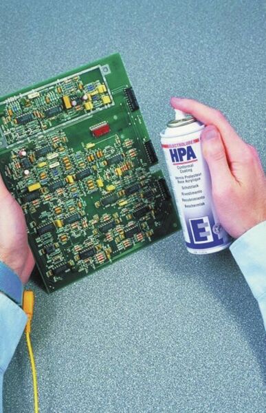 Das Bild zeigt eine Person, die eine grüne Leiterplatte in der einen Hand und eine Sprühdose mit Elektronikreiniger in der anderen Hand hält. Der Hintergrund ist grau und strukturiert.