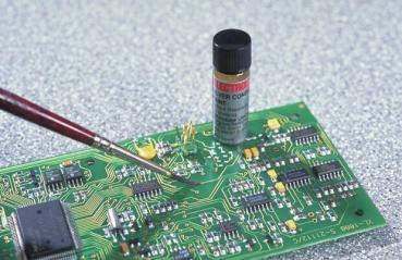 The image shows a circuit board made of green material with numerous electronic components. A red brush touches the board, while a small container stands on it.