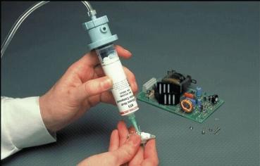 One hand is holding a syringe filled with a white material, while the other hand works on a green circuit board with various electronic components.