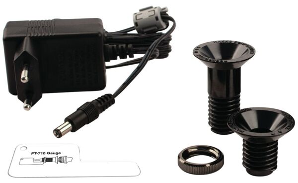 The image shows a black charger with cable and two black screw plugs. Alongside is a flat, transparent plastic measuring device.