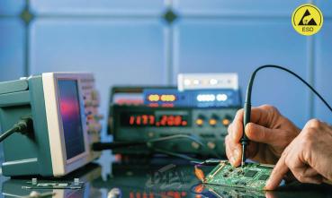 In the image, you can see a hand connecting an electronic component on a green circuit board with a testing device. In the background, there are electronic measuring instruments with various displays.
