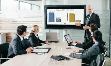 A group of five people in a modern conference room. They are sitting at a table. A man is presenting on a screen with diagrams, while the others watch attentively.