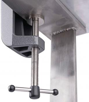 The image shows a sturdy metal clamp attached to the edge of a table. It has a long, rotating handle and a cylindrical threaded rod for fastening.