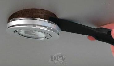Das Bild zeigt einen Handgriff, der eine runde, silberne Deckenlampe anhebt. Die Lampe hat einen transparenten Bereich in der Mitte und ist in einen dunklen, kreisrunden Rahmen eingefasst.