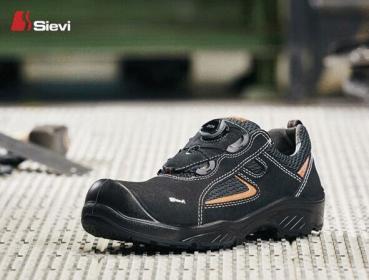 The image shows a black work shoe with a round toe cap. It has orange accents and a non-slip sole. The shoe is placed on a gray, textured surface.