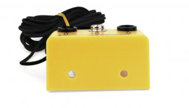 The image shows a yellow device with a rectangular shape. It has two black connectors and a screw in the middle. A black cable is attached at the back.