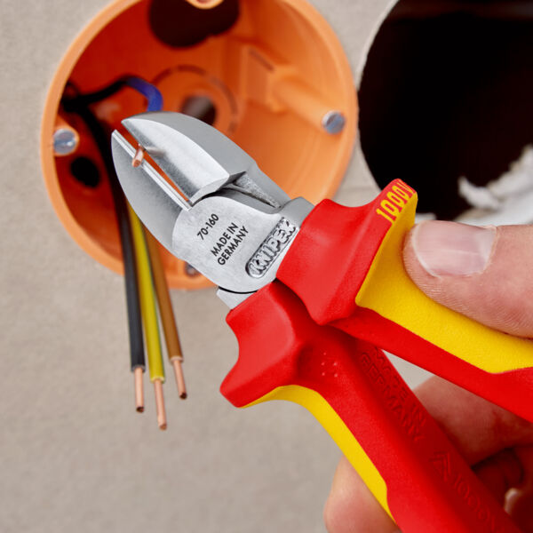 Das Bild zeigt eine Hand, die eine rote und gelbe Zange hält. Die Zange ist zum Schneiden von Drähten in der Nähe einer elektrischen Anschlussdose mit mehreren Kabeln.