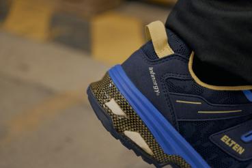 The image displays a close-up view of a sports shoe in blue and black. The shoe features yellow accents and a textured sole that provides good traction.