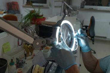 A person with blue gloves is working with a tool under a magnifying lamp. The workspace is neat and contains various tools and materials.