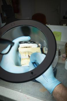A hand with blue gloves is holding a dental model under a magnifying glass. The model displays white teeth and a jaw. In the background, work tools and a blurred environment can be seen.