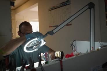 The image shows a person wearing gloves, working intently at a table under a bright, round work lamp. In the background, a wall with a calendar is visible.