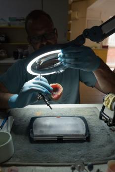 A person wearing glasses and gloves is working under a lamp. They are holding a model of a denture in their hand while working on it with a tool. The background is blurred.