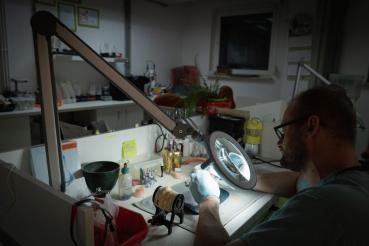 A man works intently at a table under a large, illuminated magnifying glass. Around him are tools and materials that create a workshop atmosphere.