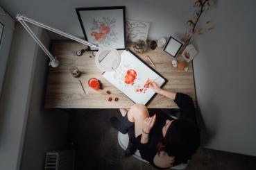 The image shows a table with art supplies. A person is sitting there, painting with reddish colors. Around the table, there are several objects, including a frame and a plant.