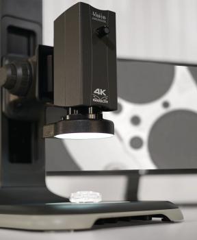 Das Bild zeigt ein Mikroskop mit einer schwarzen Linse, das auf eine kleine, transparente Probe gerichtet ist. Im Hintergrund sieht man eine grafische Darstellung, die an Film erinnert.