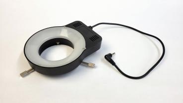 Das Bild zeigt einen runden Lichtring mit einem schwarzen Gehäuse, das an einer Seite mit einem Kabel verbunden ist. Der Innenbereich ist hell, der Ring hat zwei seitliche Halterungen.
